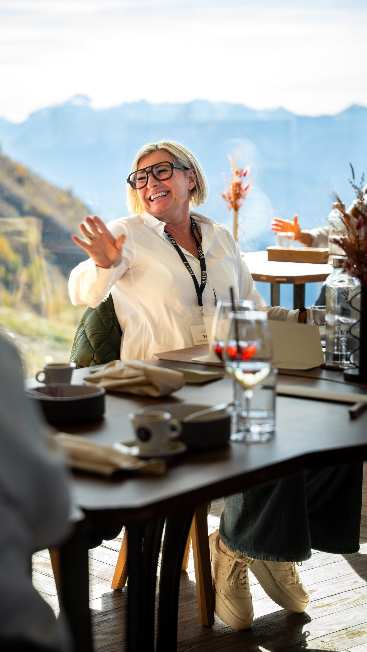 Sportwagen Erlebnisreisen: Fröhliche Teilnehmerin beim Gourmet-Essen mit Blick auf das Alpenpanorama.